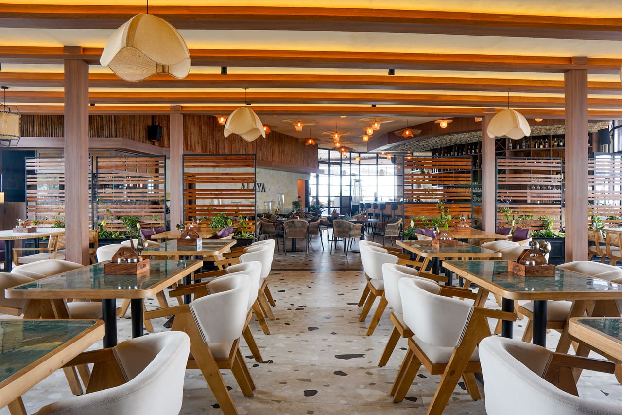 The Ālaya dining hall by day: a central aisle of green marble tables and cream chairs under timber beams and fabric pendants, with the bar and windows beyond.