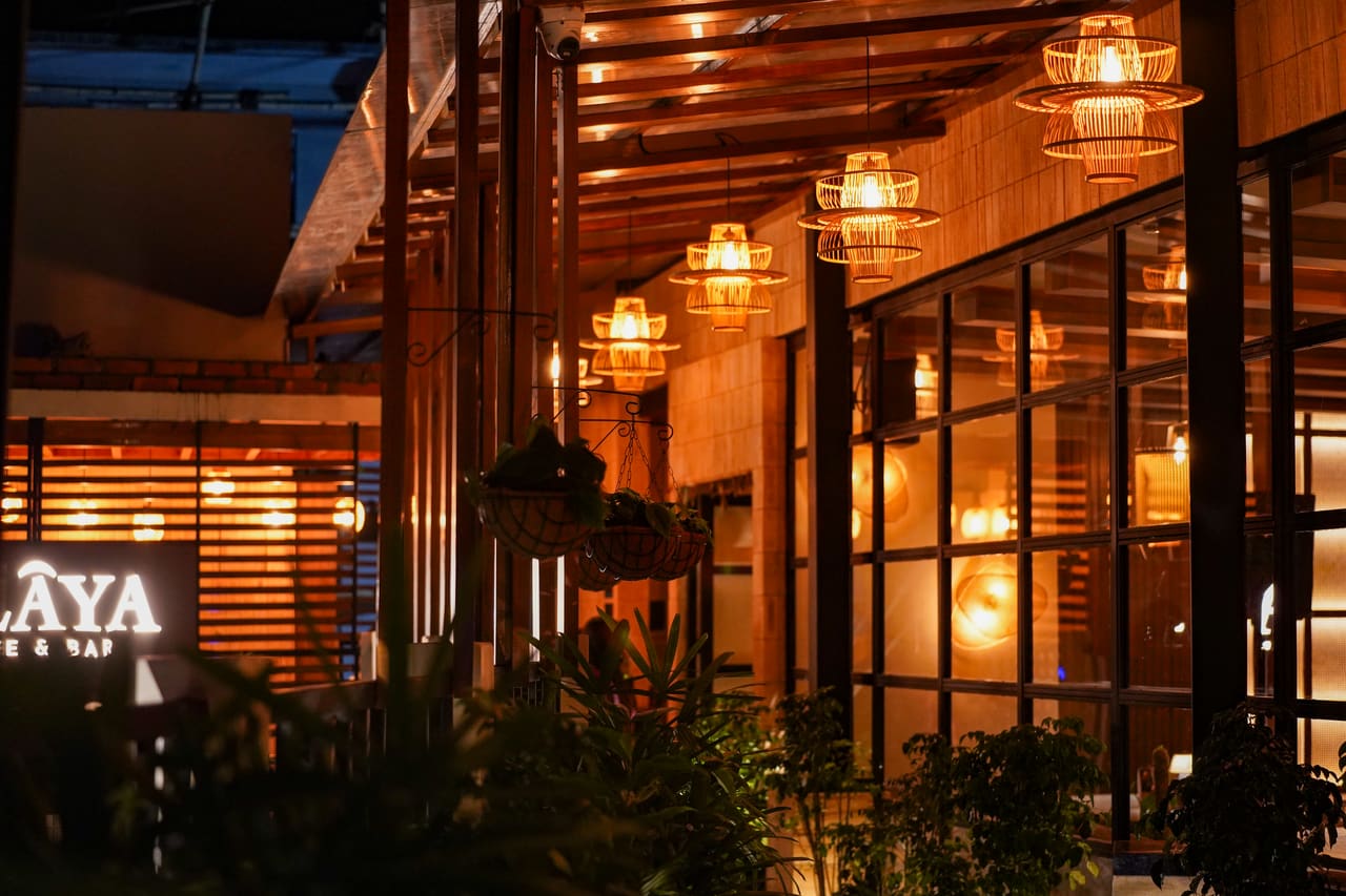 Ālaya’s glazed frontage at night, rattan pendants lit along the timber canopy and planters below.