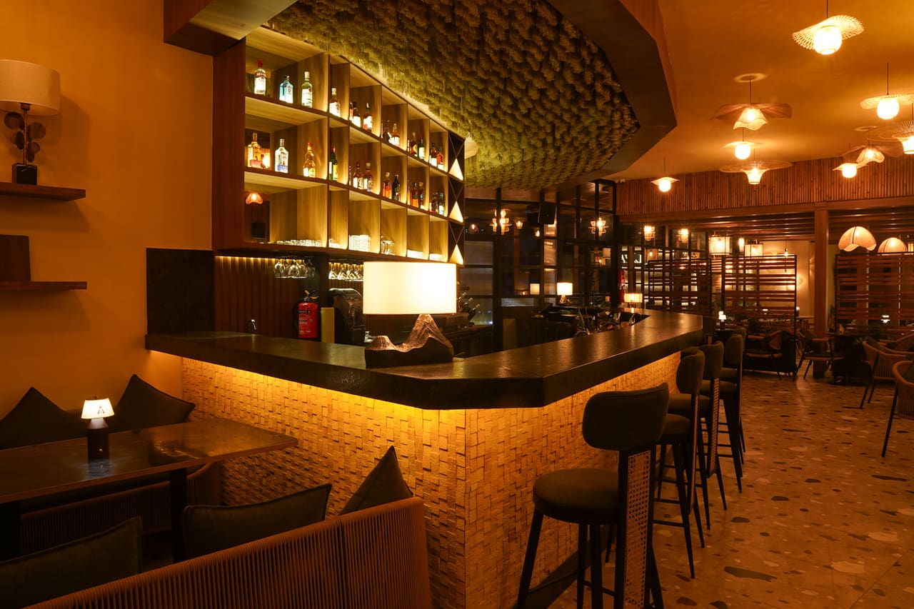 The curved Ālaya bar at night: a lit counter with tall stools beneath backlit bottle shelving and a woven ceiling panel.