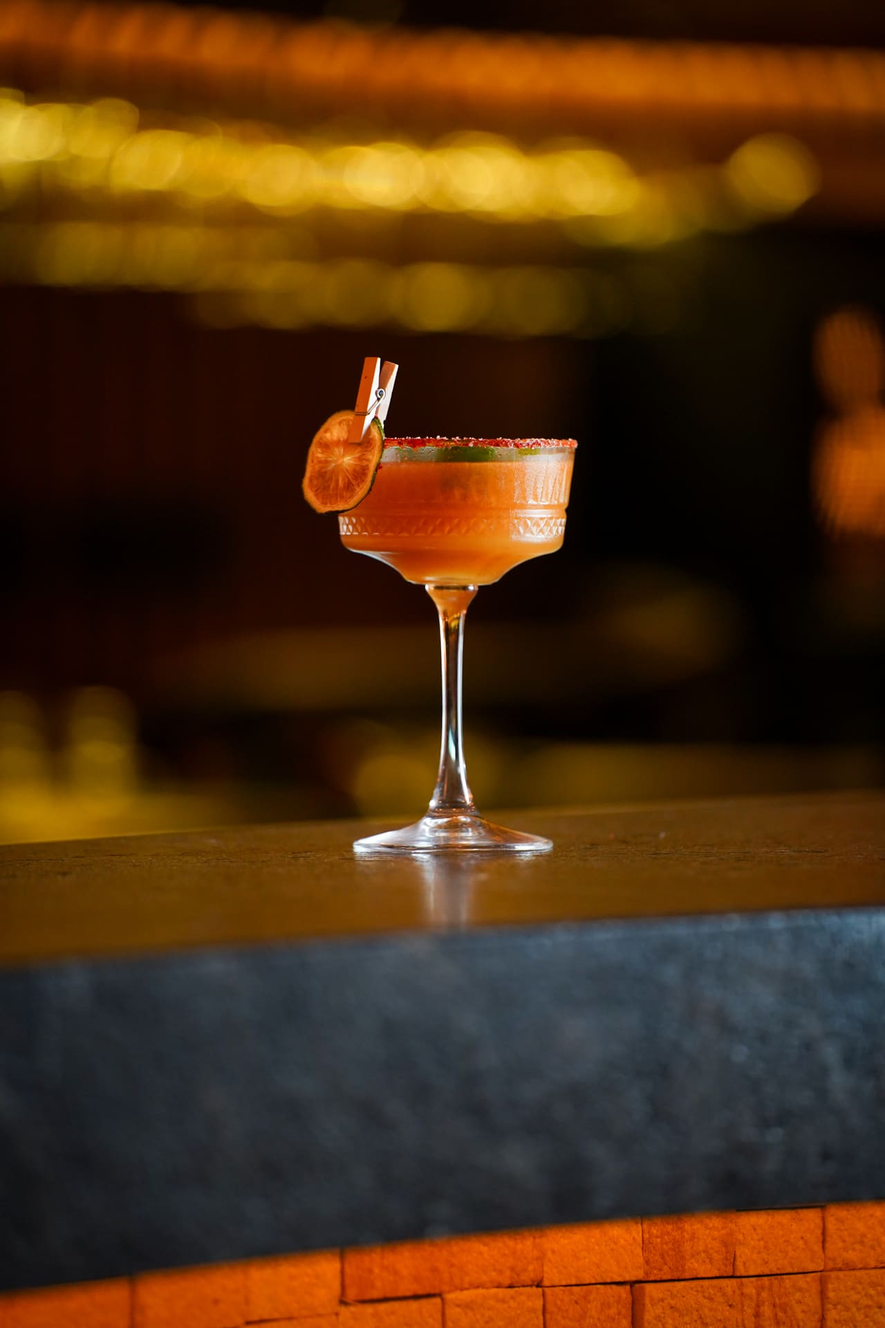A coral cocktail with a dried citrus garnish in a coupe glass on a stone bar top, warm lights out of focus behind.