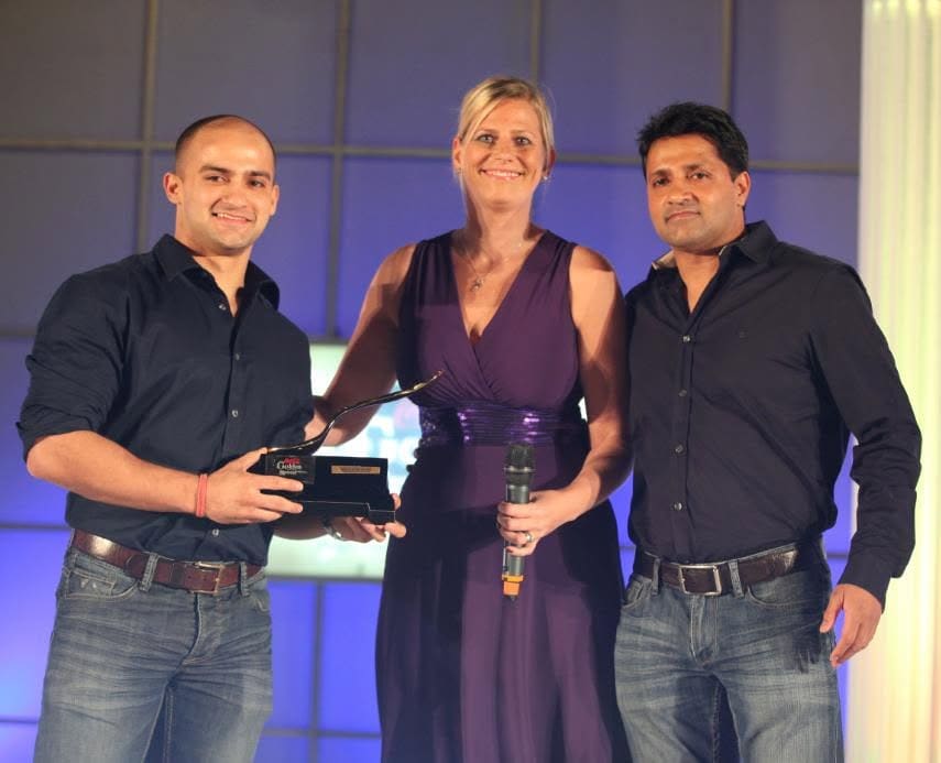 Rish Saini (left) and his father Preet Saini (right) on stage holding an award, with a presenter between them.