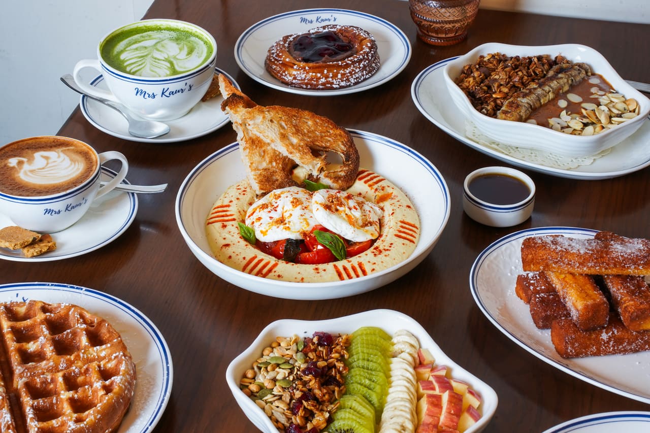 A breakfast spread on a timber table: eggs, waffles, an açaí bowl, French toast and lattes in branded cups.