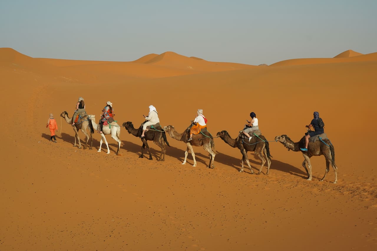 A camel caravan crossing orange sand dunes under a hazy sky, Morocco.