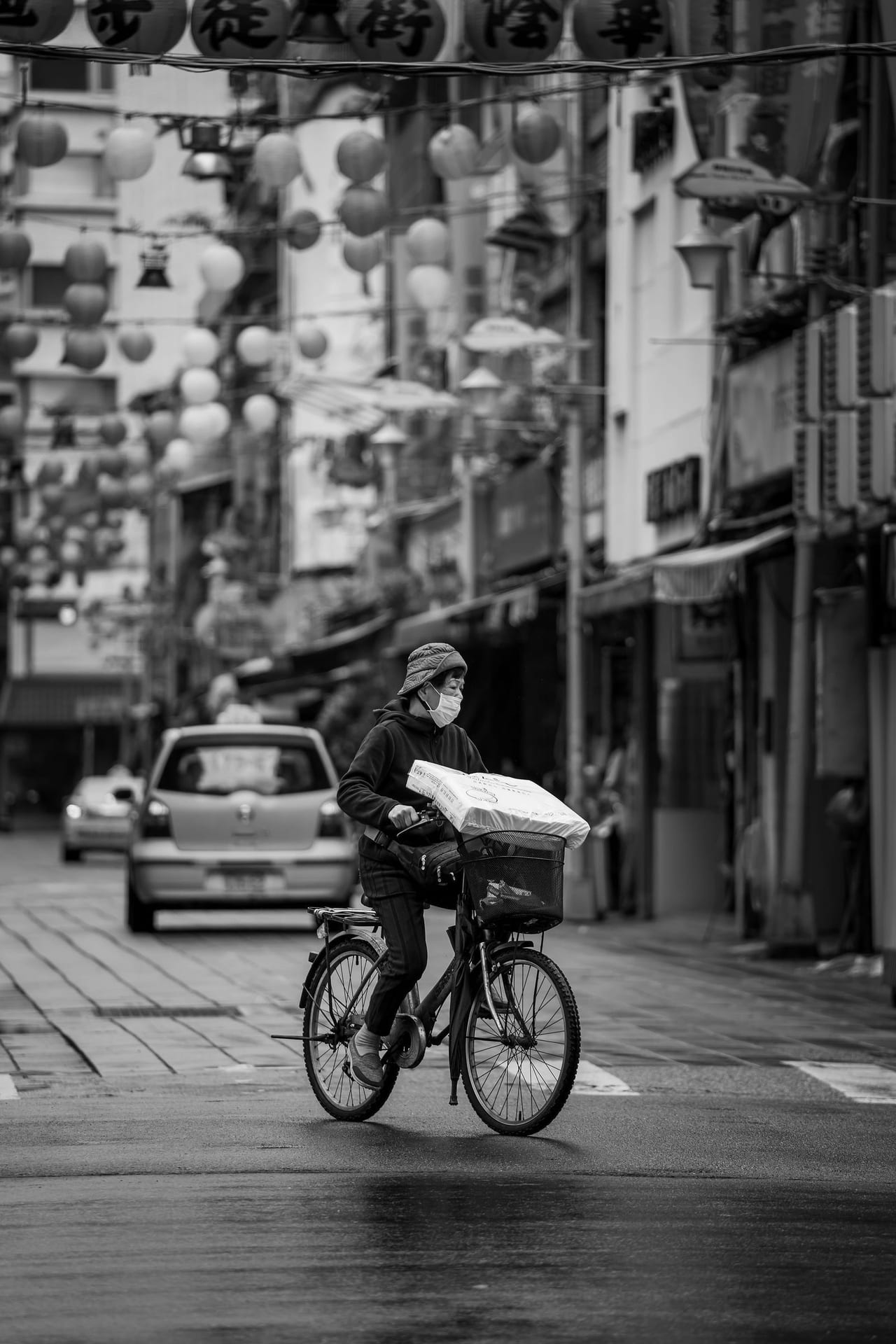 A cyclist on a narrow city street lined with signs and shopfronts, in black and white