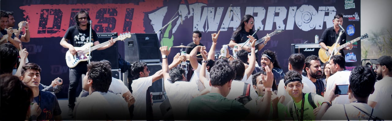 A band playing on the festival stage under a large Desi Warrior backdrop, the crowd in front