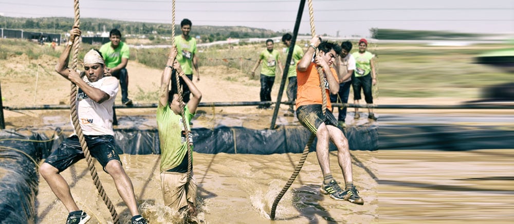 Runners swinging on ropes over a muddy water pit on the Mowgli obstacle