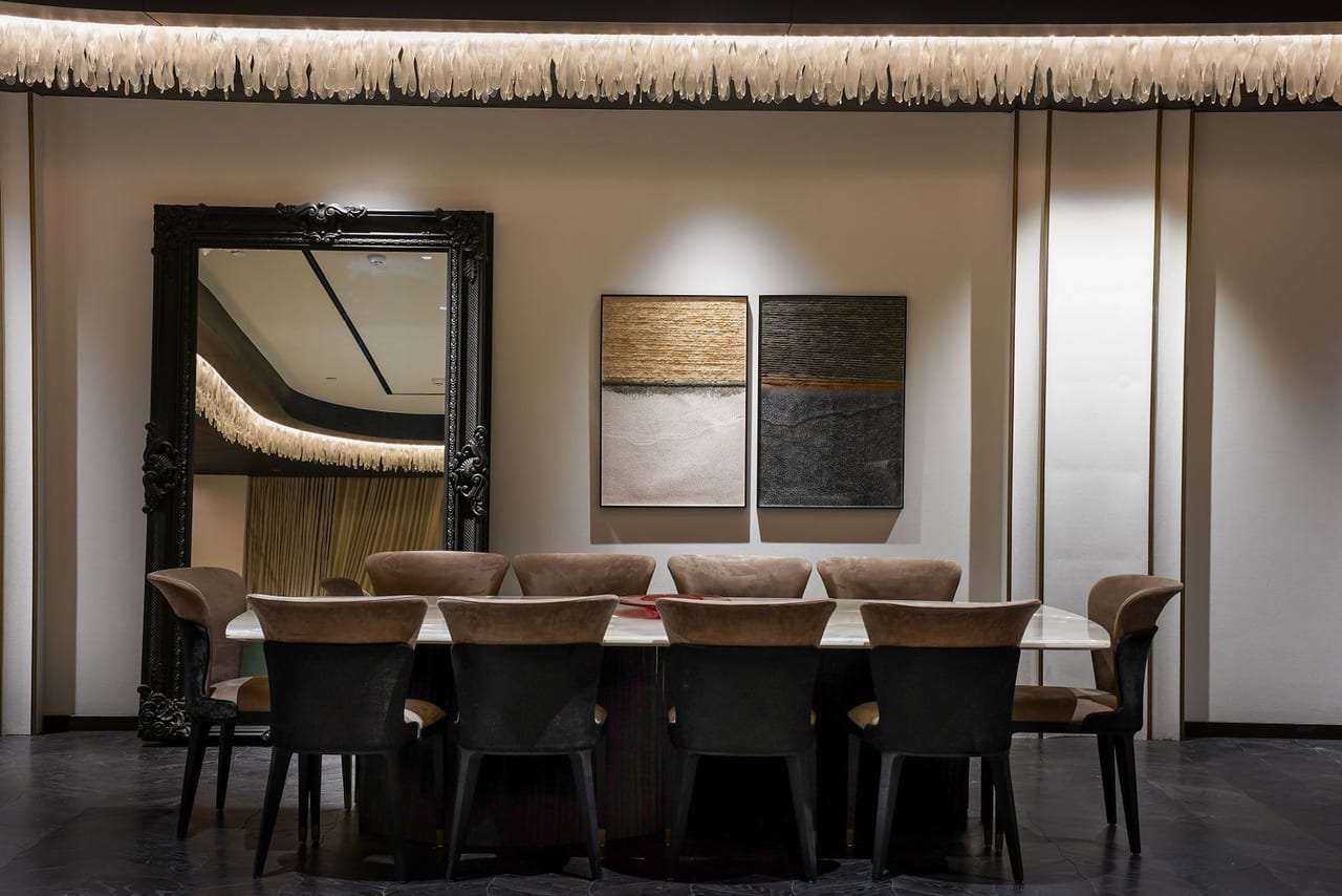A dining room with a long table, an ornate black mirror and two textured artworks under a fringed light cove