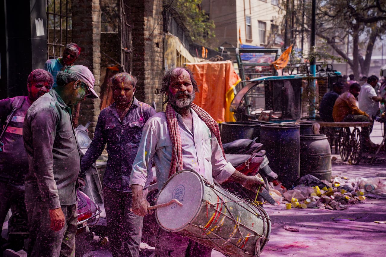 A drummer playing a dhol leads people through a street during Holi, purple powder on the ground.
