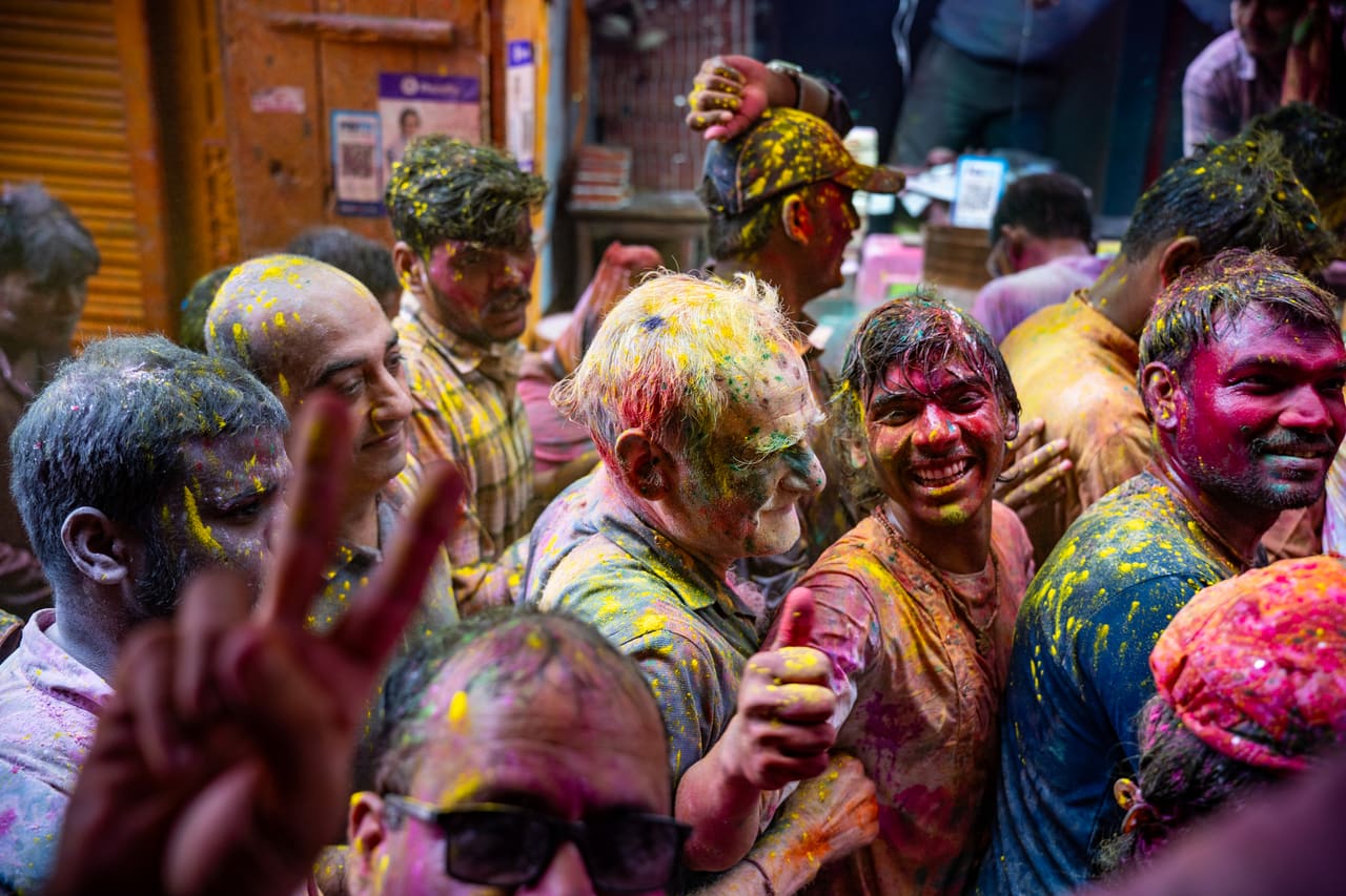 Holi revellers covered in coloured powder crowd together, one smiling man making a victory sign.