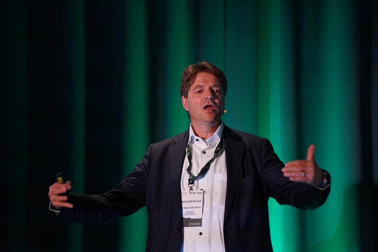 A keynote speaker with arms open under green stage light at the MEMS World Summit, Porto
