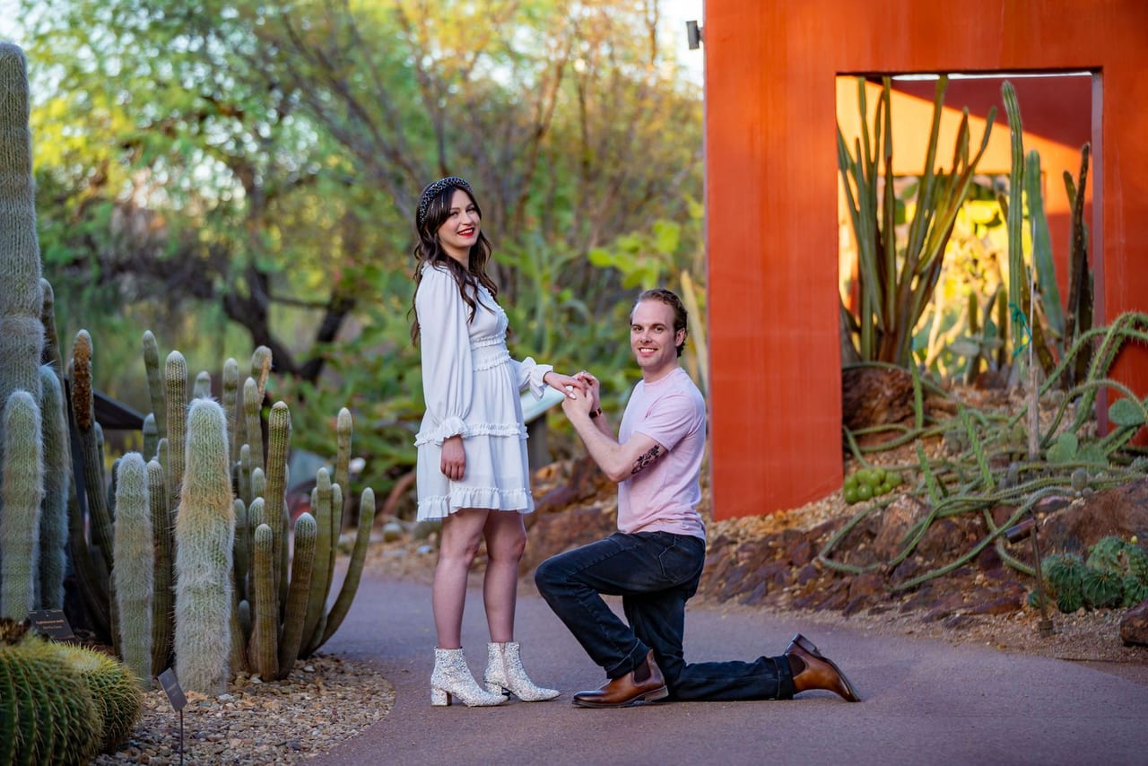 A man on one knee proposing beside a bright orange wall among cacti