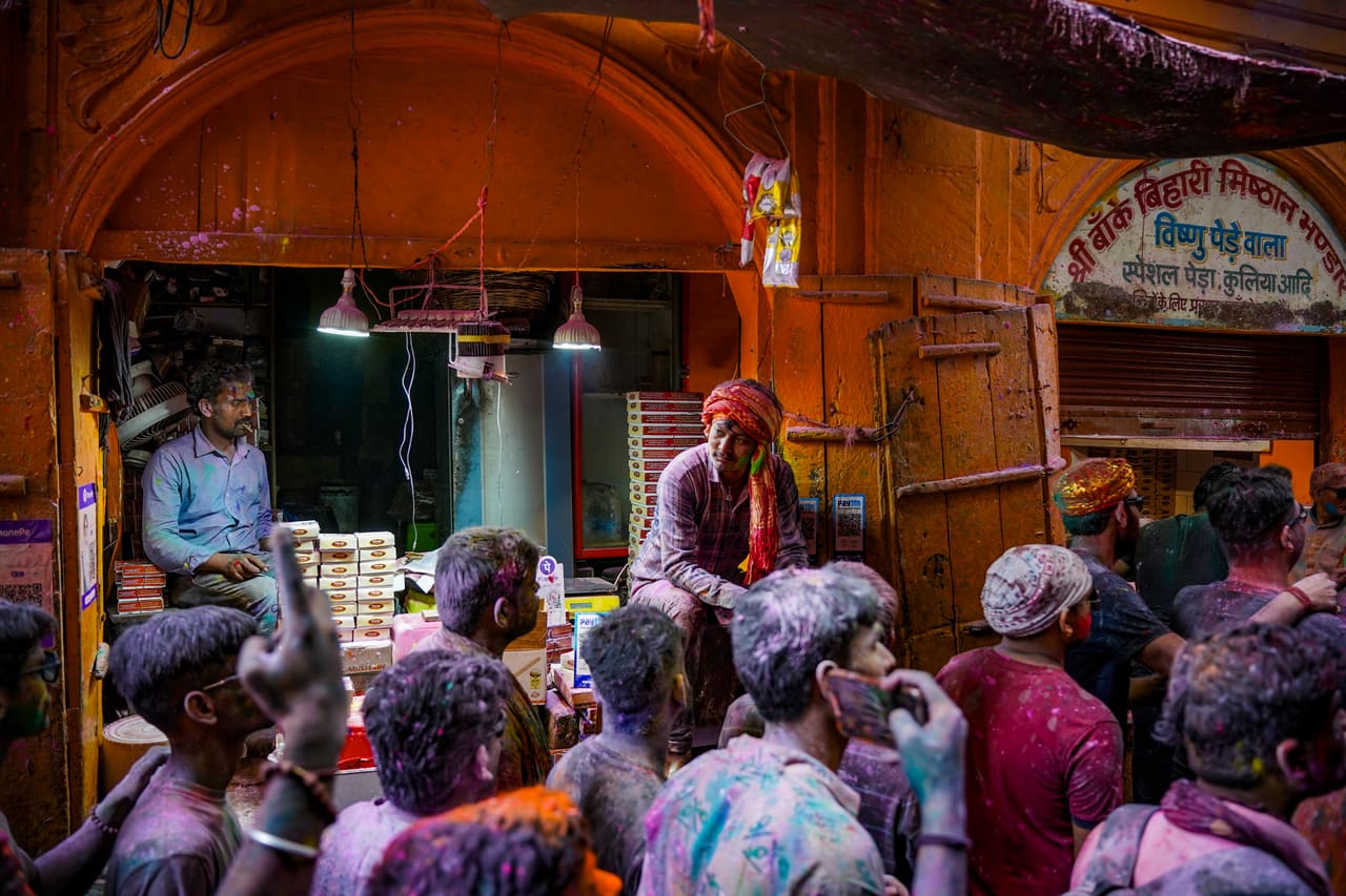Men covered in Holi colour gather in front of an old sweet shop with a painted Hindi sign.