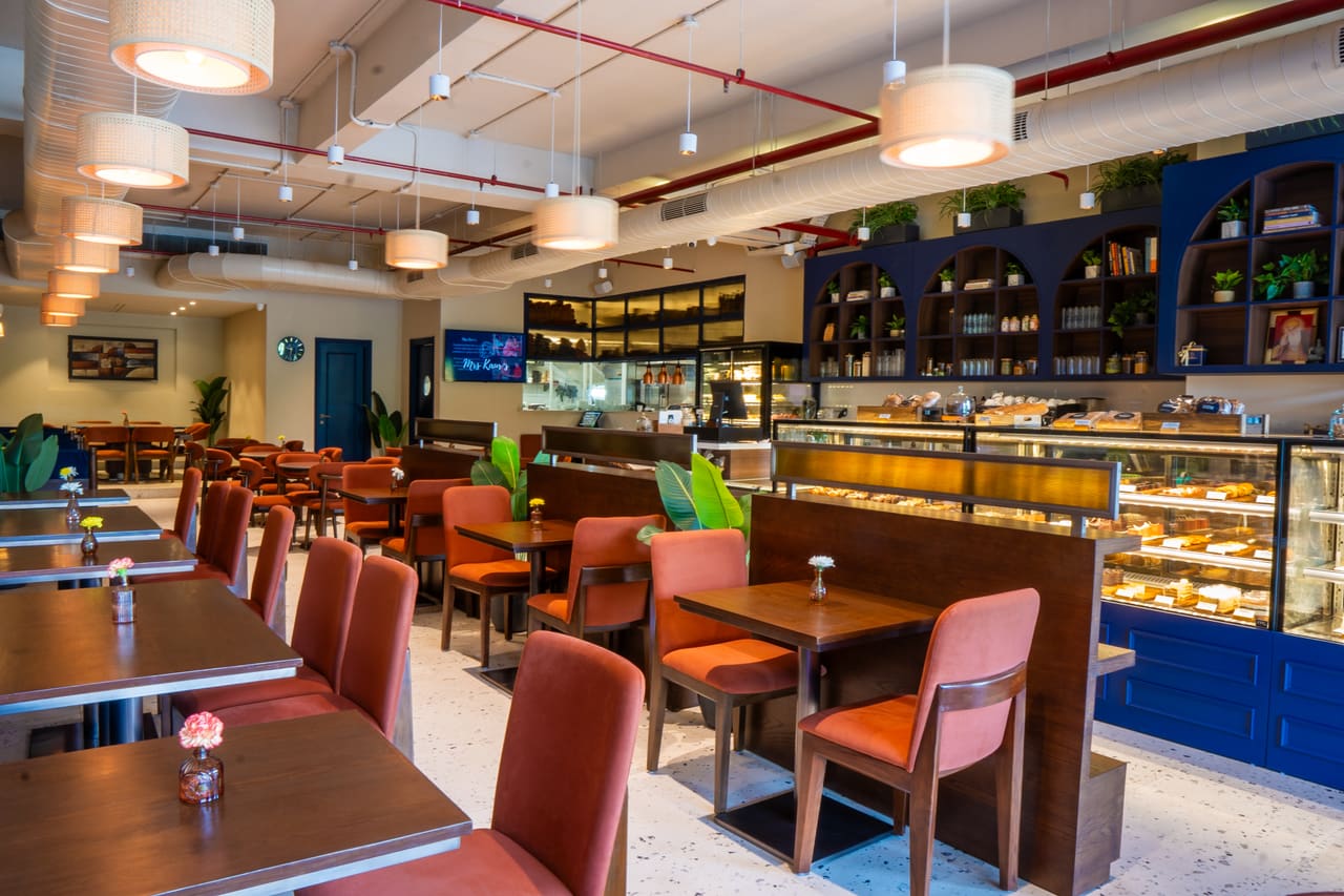 The main dining room: orange upholstered chairs at timber tables under drum pendants, with the bakery counter along the right.