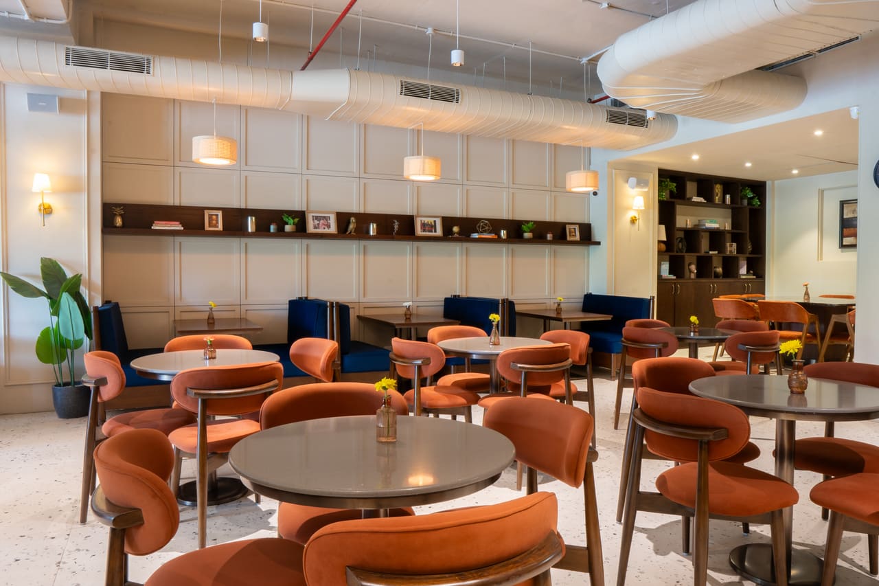 A bright seating area with round tables, curved orange chairs, wall shelves and drum pendants.