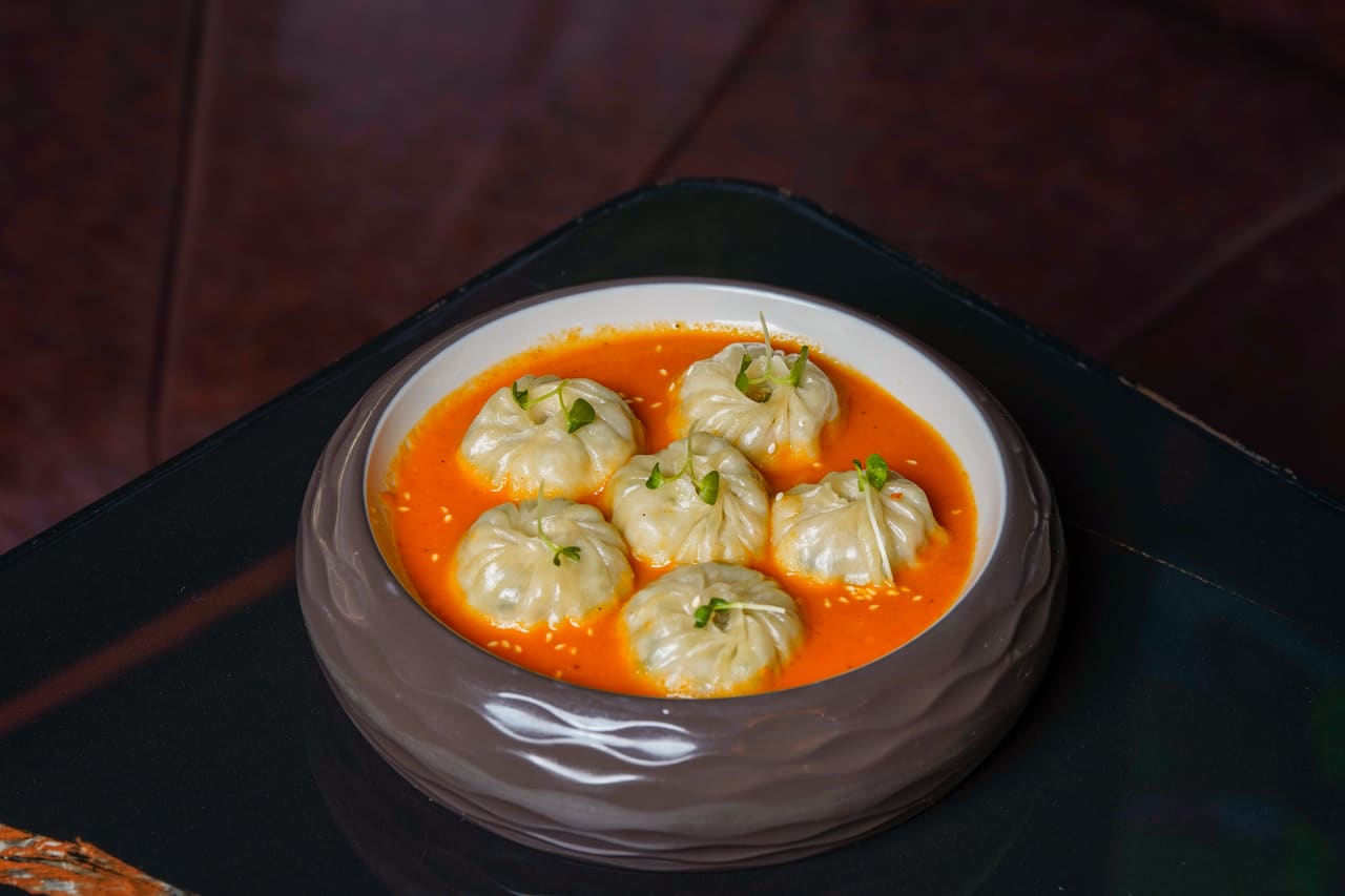 Momos in a shallow bowl of orange curry sauce on a dark surface.