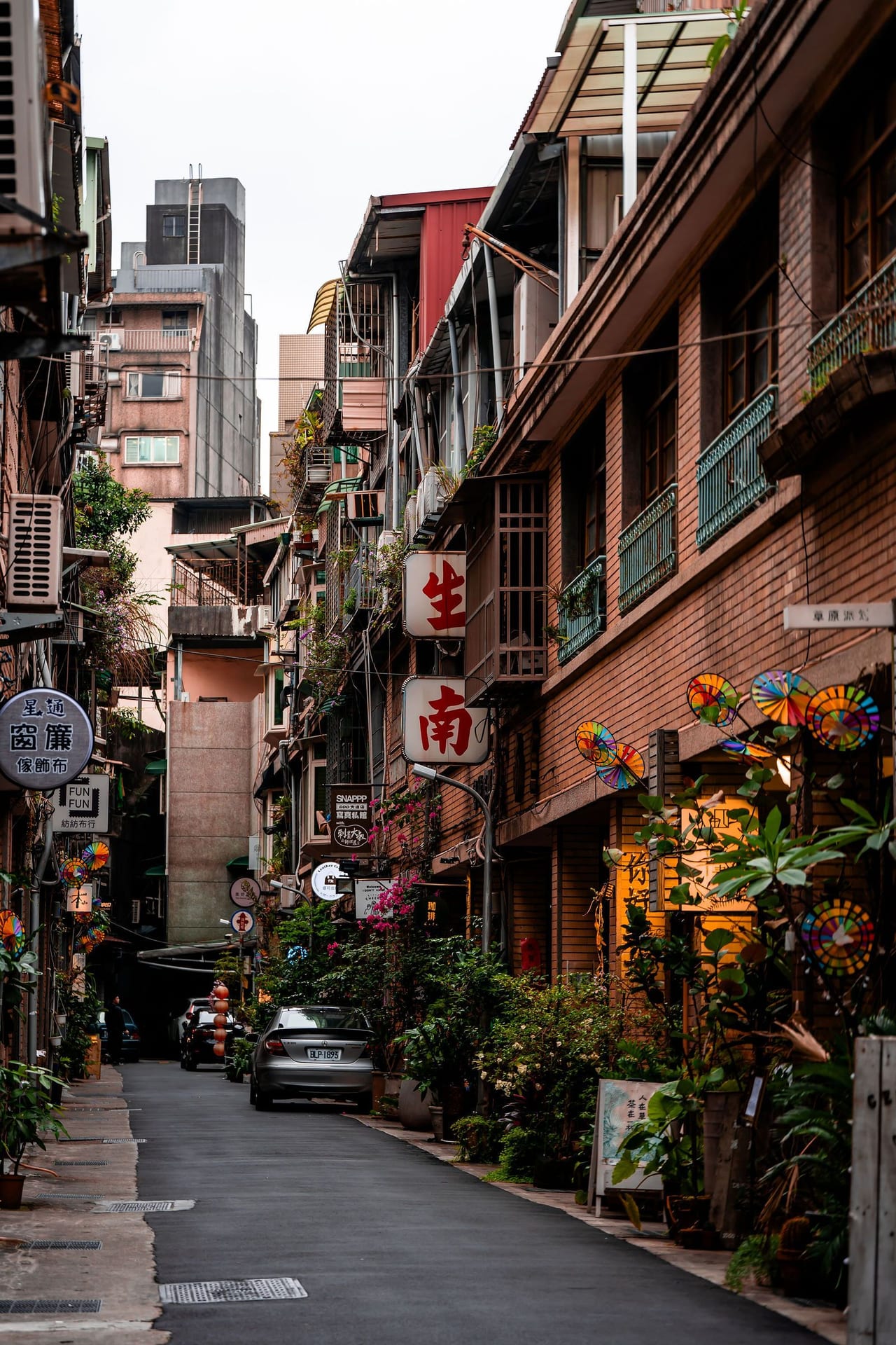 A narrow lane between older apartment buildings, with plants and lanterns along it