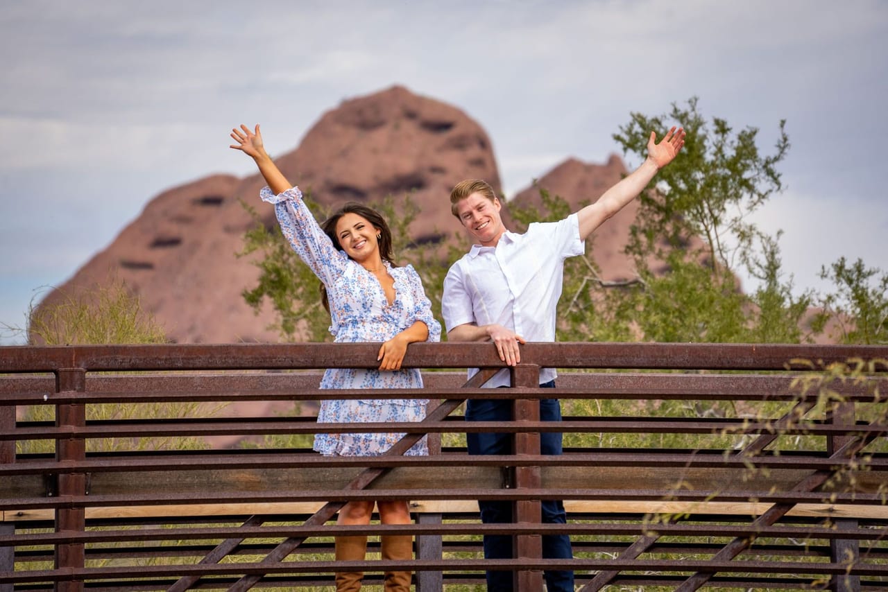 A newly engaged couple cheering with their arms raised on a wooden bridge below red rock buttes