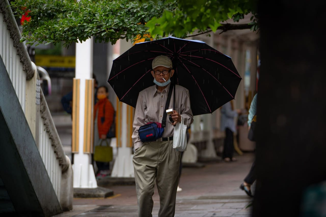 An older man with a black umbrella and a shoulder bag on a city pavement