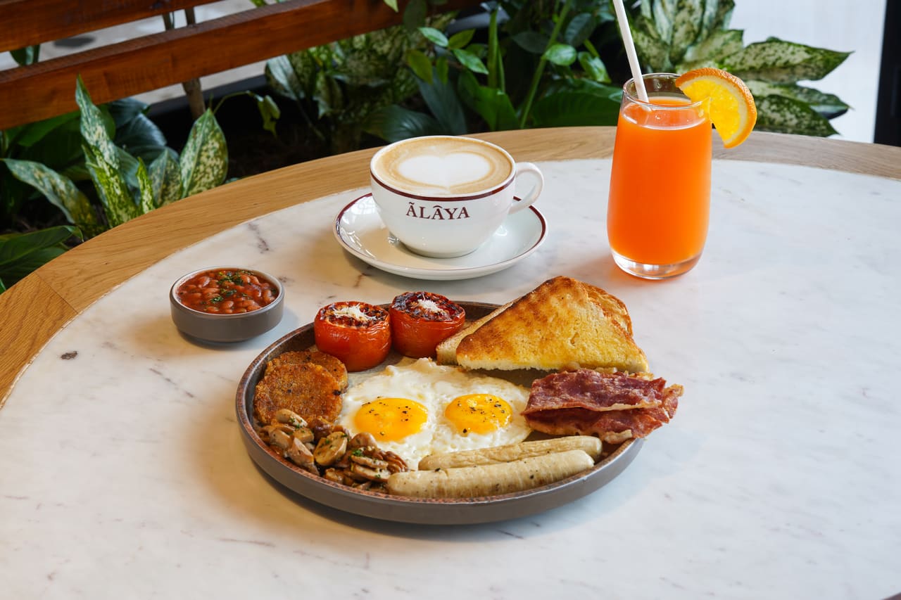 A full breakfast on a white plate beside a latte in an Ālaya-printed cup and a glass of orange juice.