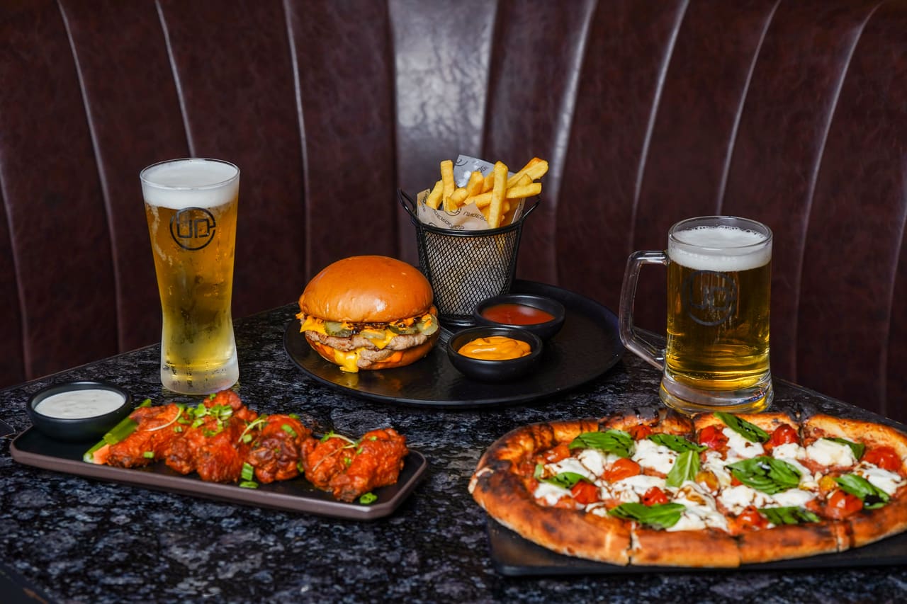 Pizza, burgers, fried chicken and pints of beer arranged on a dark stone table against a leather banquette.