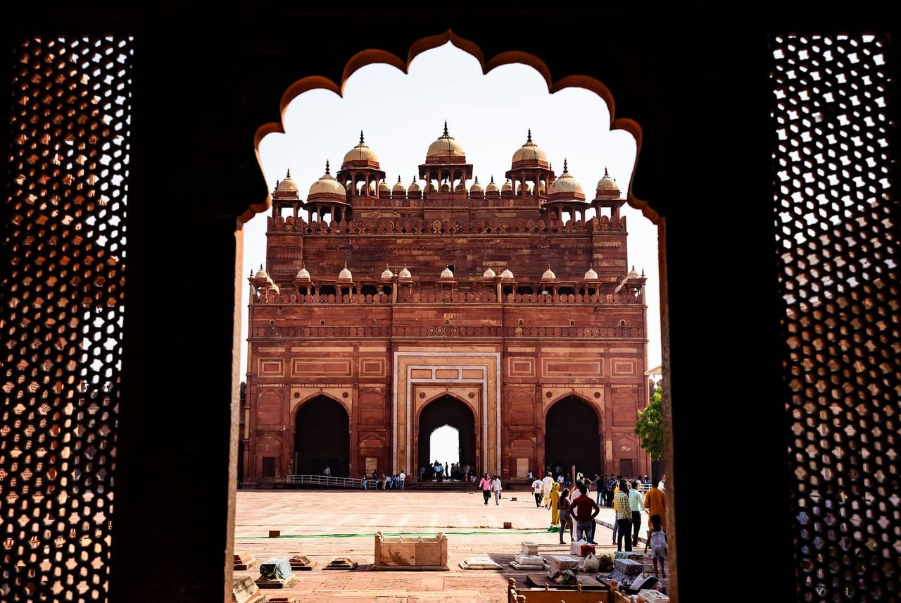 A red sandstone gateway with white domes, seen through a carved stone screen and arch