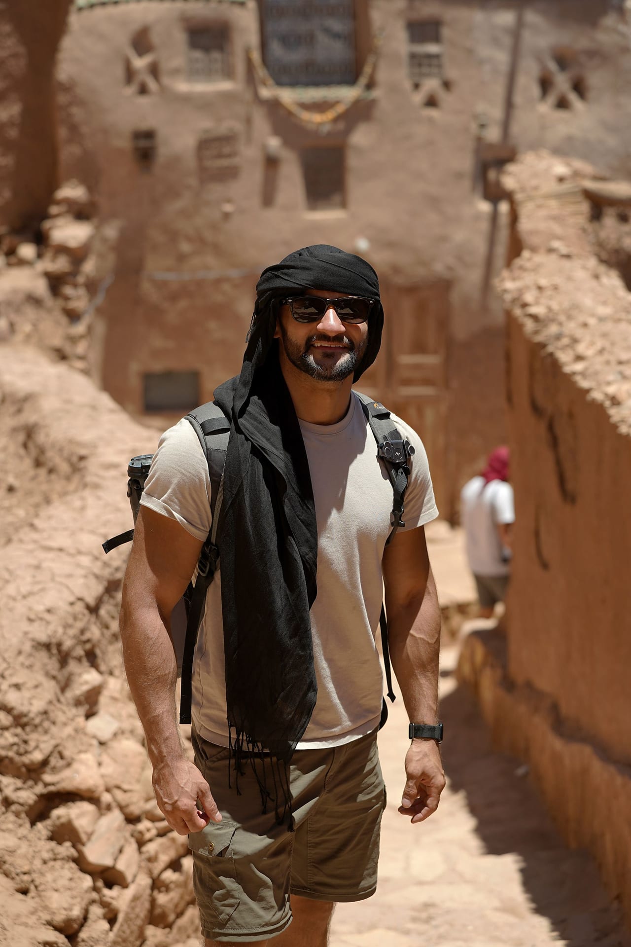 Rish Saini walking up a narrow earthen lane in Aït Benhaddou, in a headscarf and sunglasses, smiling