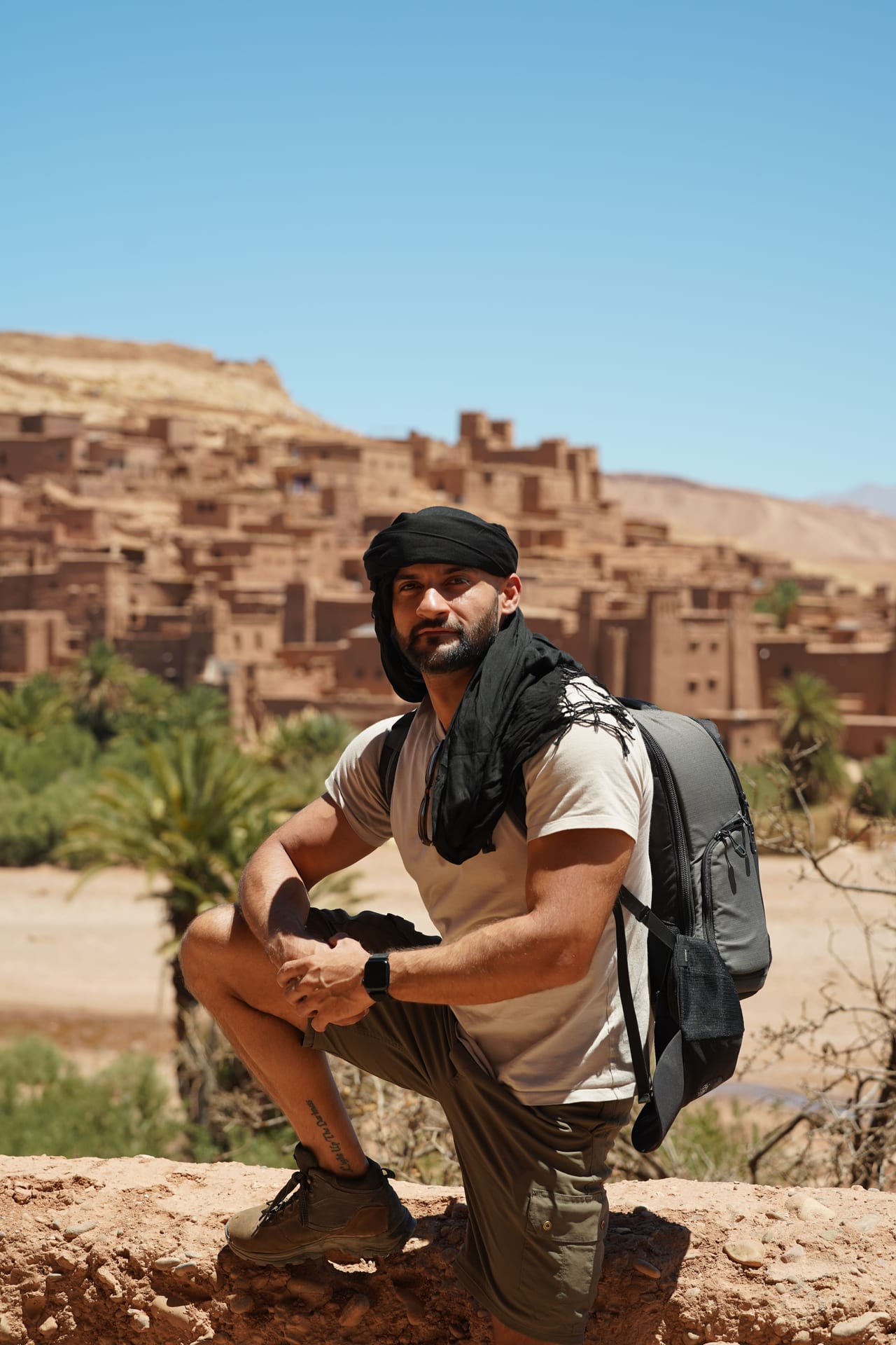 Rish Saini crouching on a rock above the earthen kasbah of Aït Benhaddou, in a black headscarf and sunglasses, with a backpack