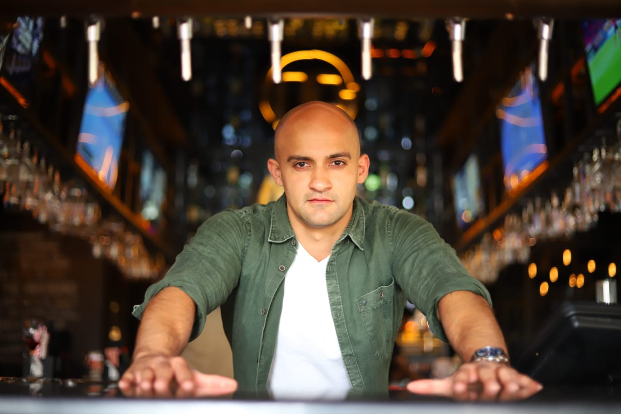 Rish Saini leaning on a bar counter with both hands, smiling, in a green shirt over a white T-shirt, warm lights behind