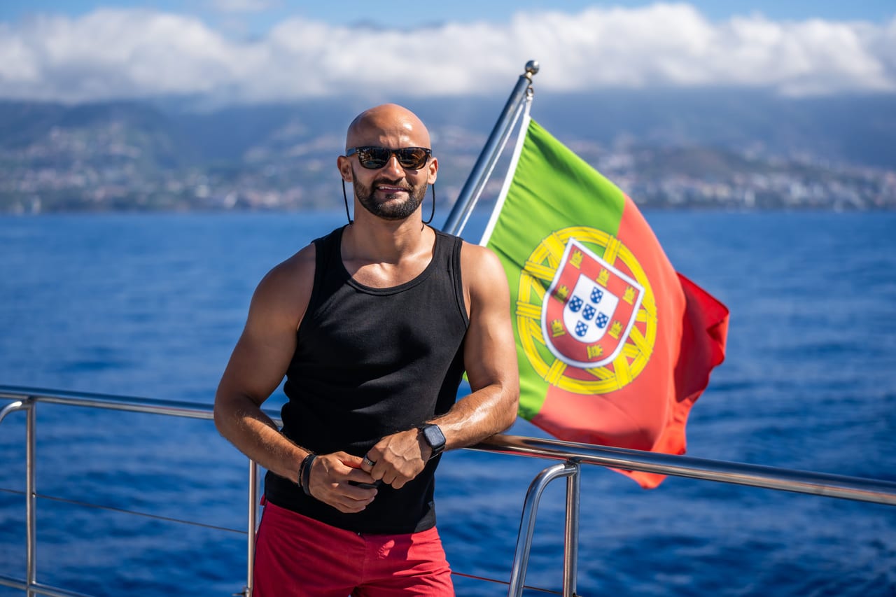 Rish Saini smiling on the deck of a boat beside a Portuguese flag, with the sea and a mountainous coastline behind