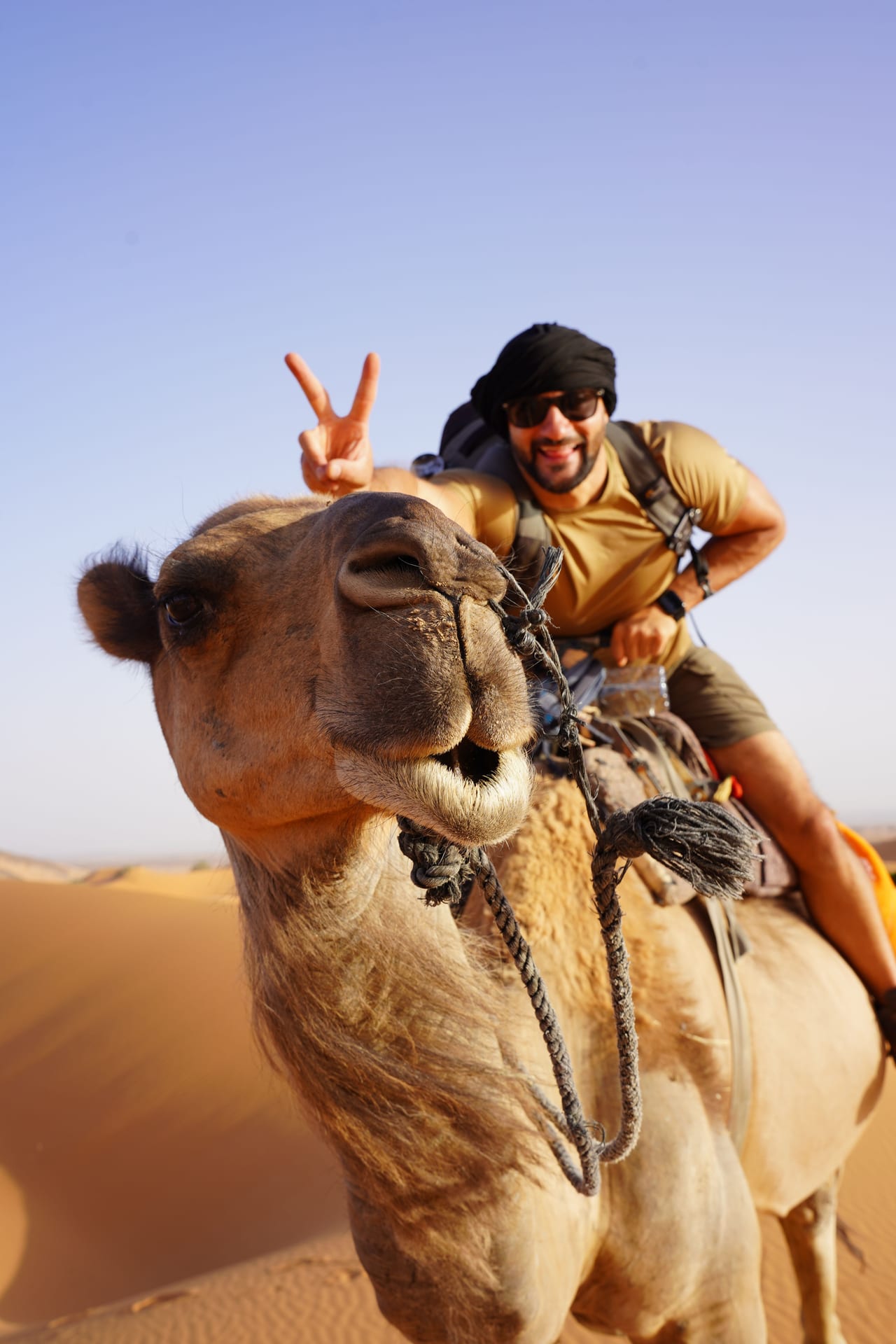 Rish Saini on a camel in the dunes, making a peace sign behind the camel’s head