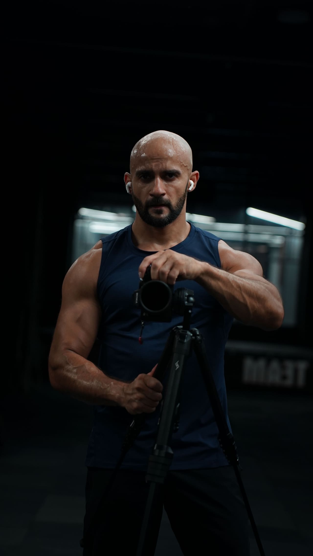Rish Saini in a sleeveless top, standing behind a camera on a tripod, lit against a dark background.