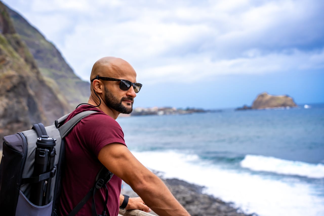 Rish Saini in sunglasses with a grey backpack, looking out over a rocky shoreline and waves