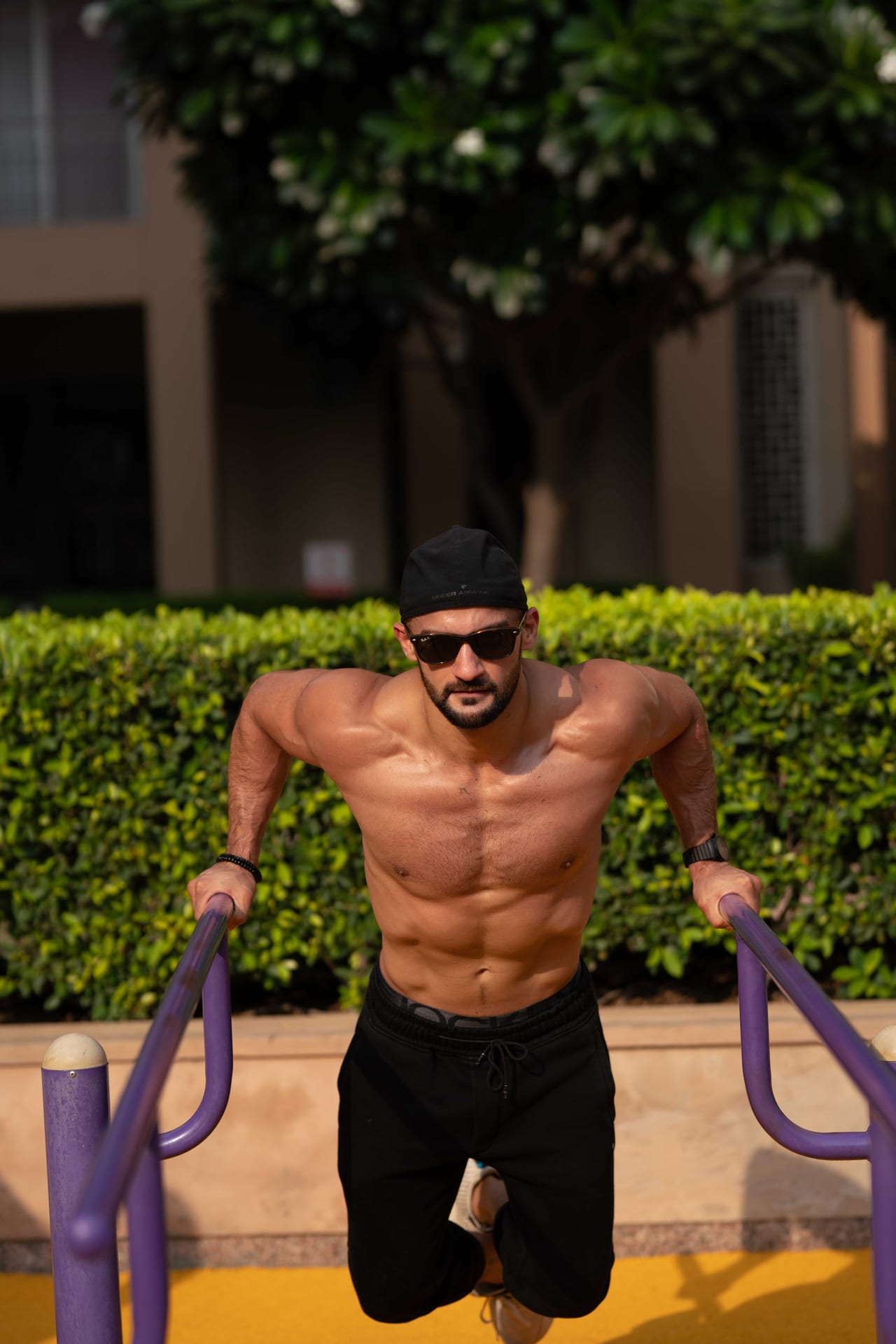 Rish Saini holding himself up on parallel bars at an outdoor gym, in sunglasses and a black cap