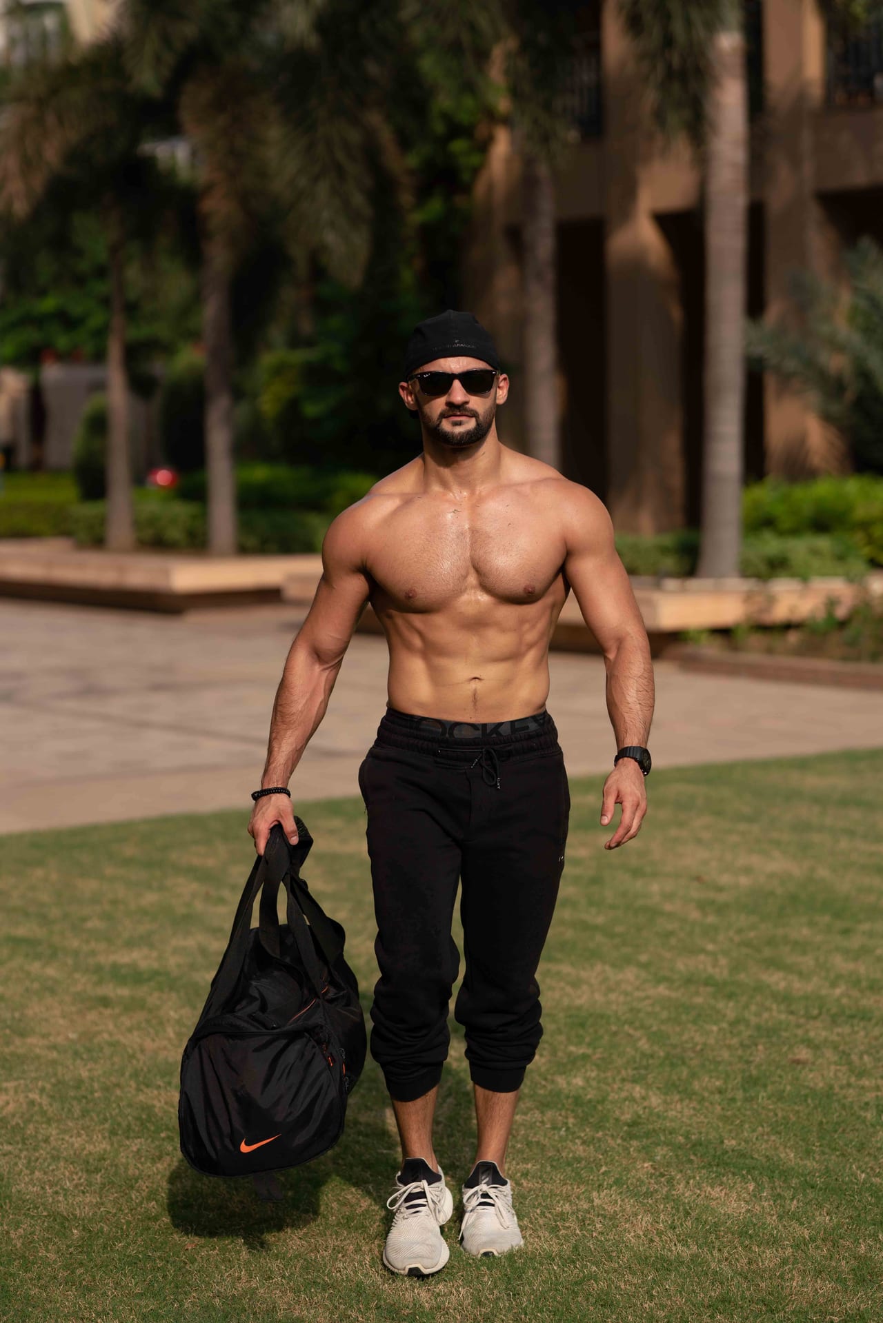 Rish Saini walking across a lawn with a gym bag in one hand, in sunglasses and a black cap