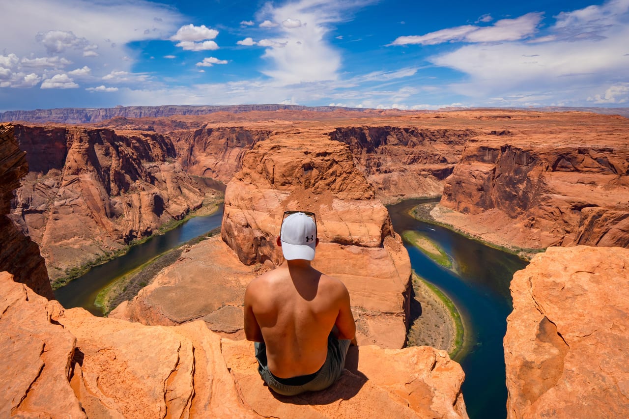 Rish Saini, seen from behind in a white cap, sitting on the sandstone rim above the river’s curve at Horseshoe Bend