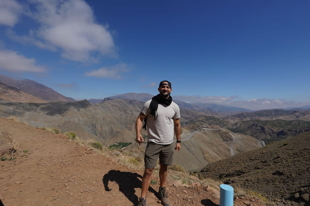 Rish Saini standing on a dry hillside above a wide mountain valley under a blue sky