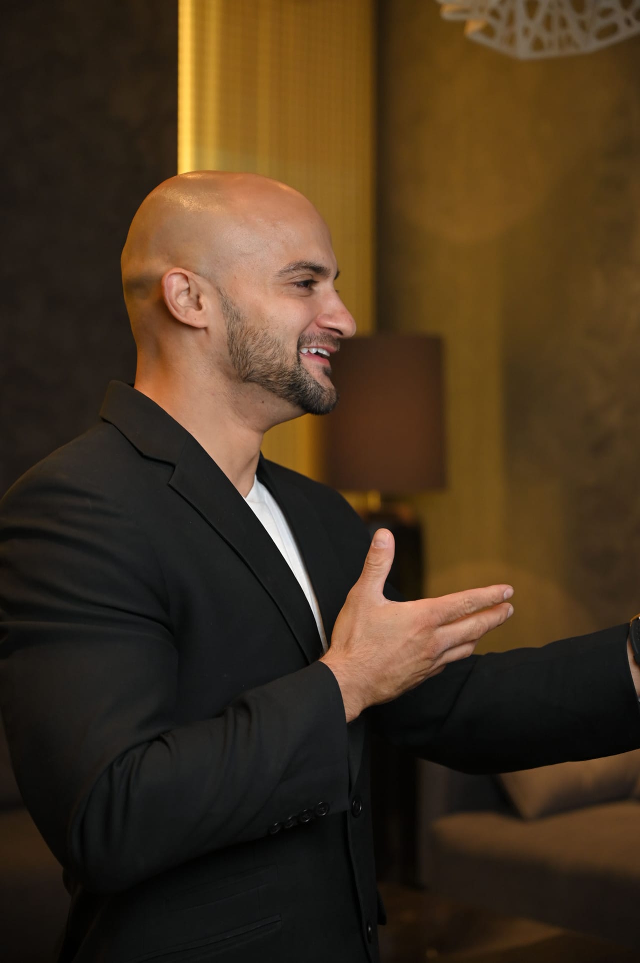 Rish Saini in a black blazer, mid-conversation, gesturing with one hand in a warmly lit room