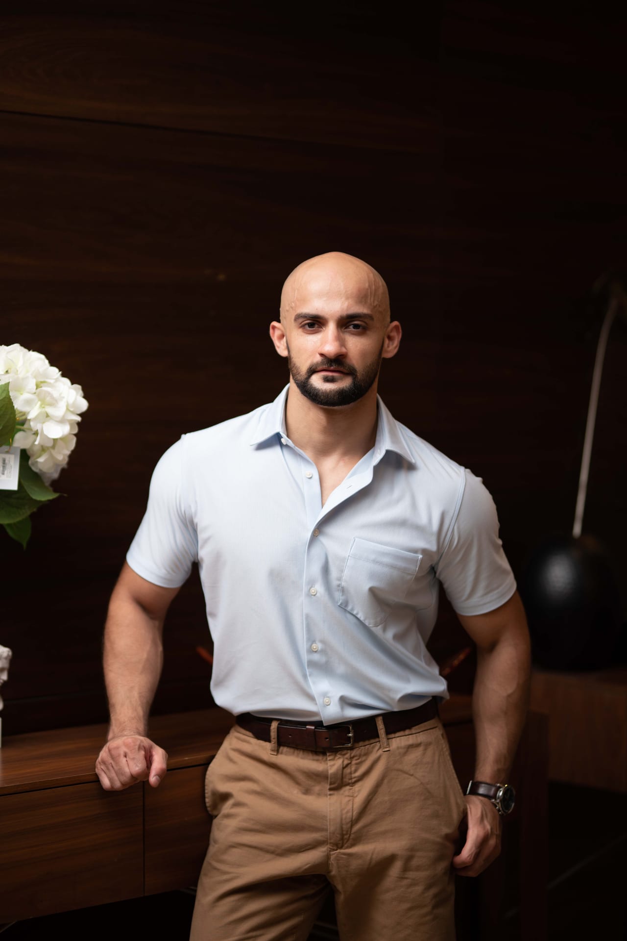 Rish Saini standing in a light blue shirt and tan trousers in a dark wood-panelled room