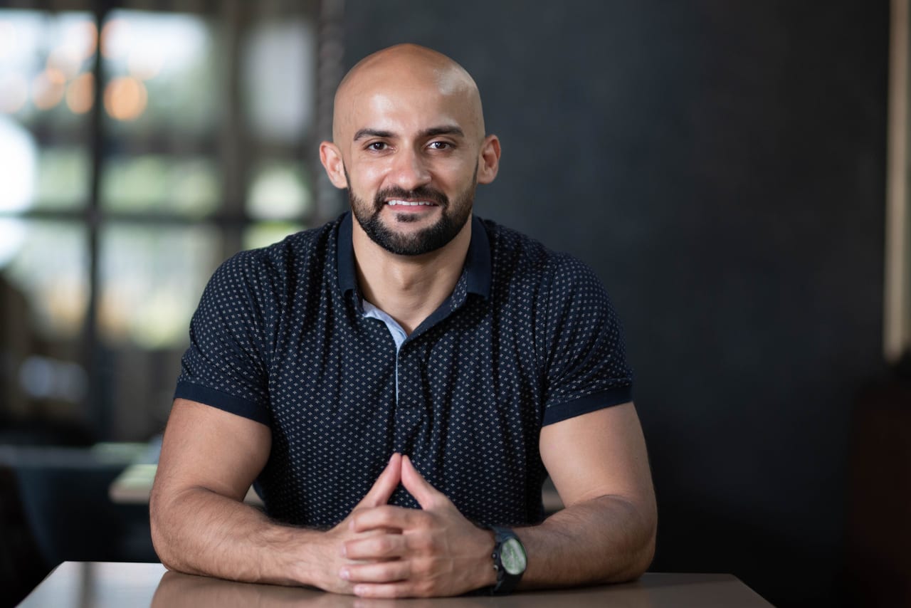 Rish Saini in a navy polo shirt, seated at a table with his hands together, smiling