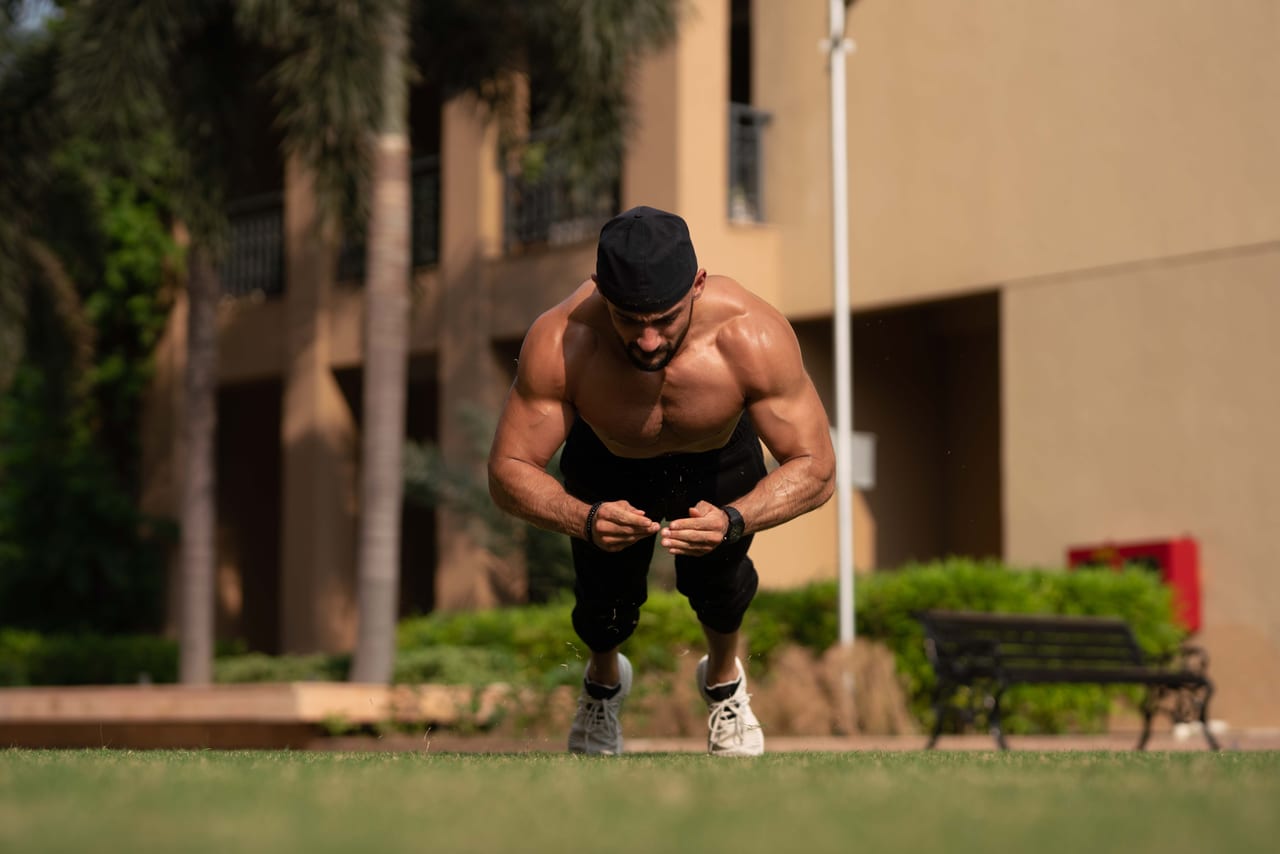 Rish Saini doing a push-up on a lawn outside a building, in a black cap