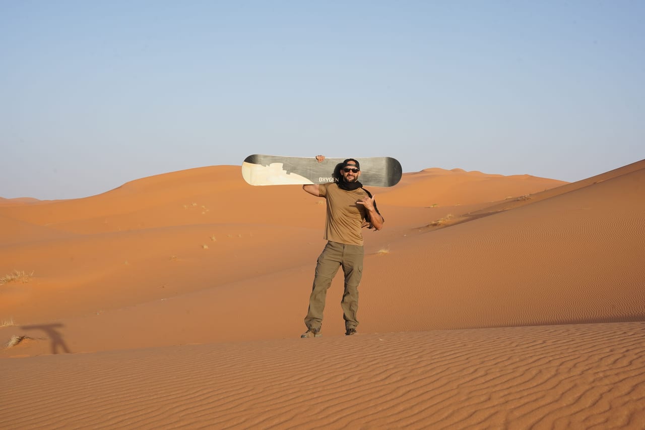 Rish Saini carrying a sandboard on his shoulder across the orange dunes