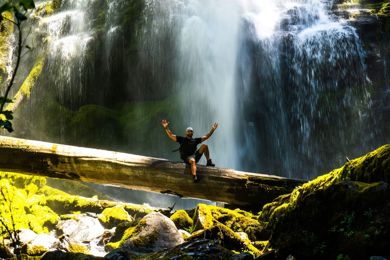 Rish Saini raising his arms on a fallen log in front of a tall waterfall and bright green moss