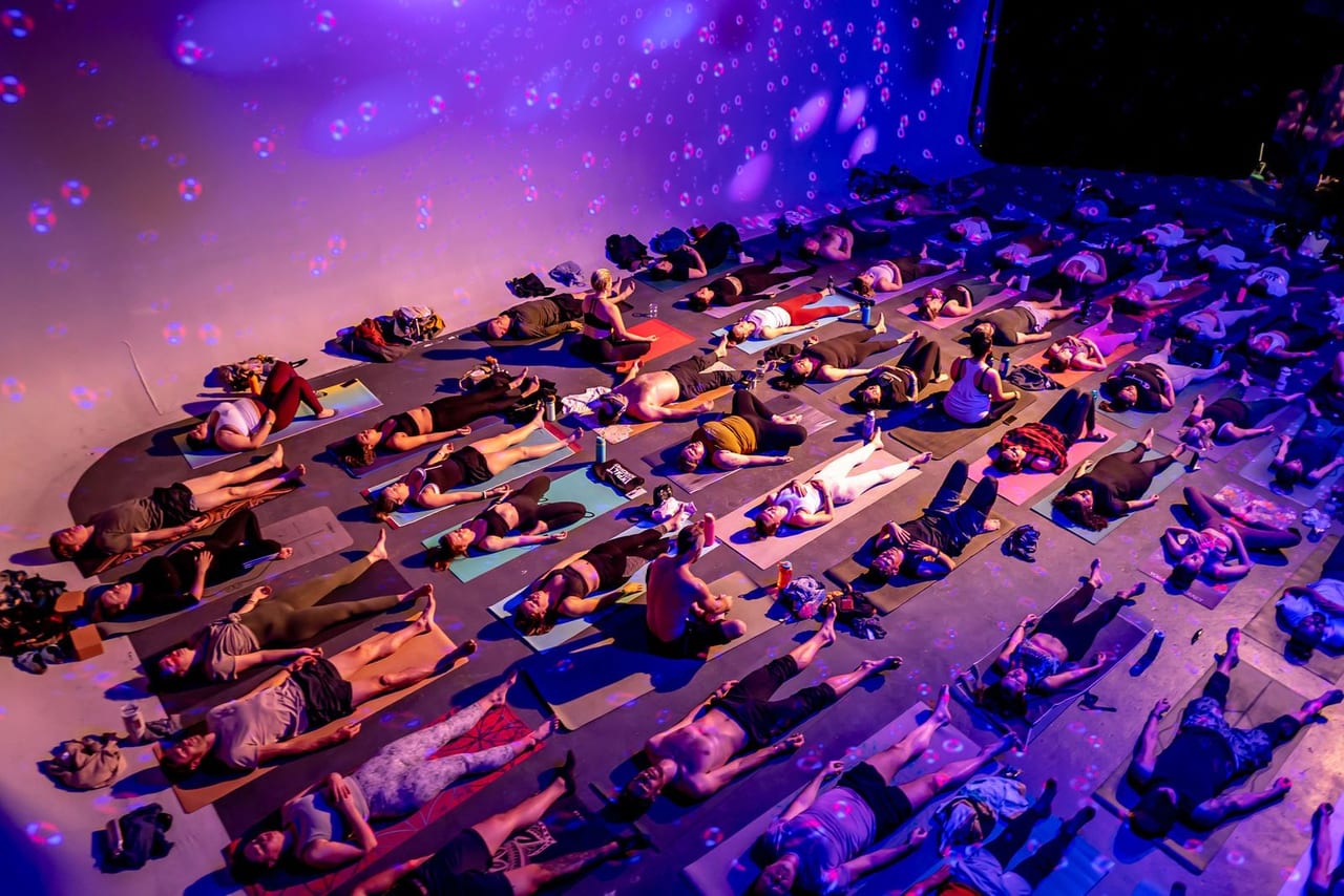 Rows of people lying on yoga mats under purple light, seen from above