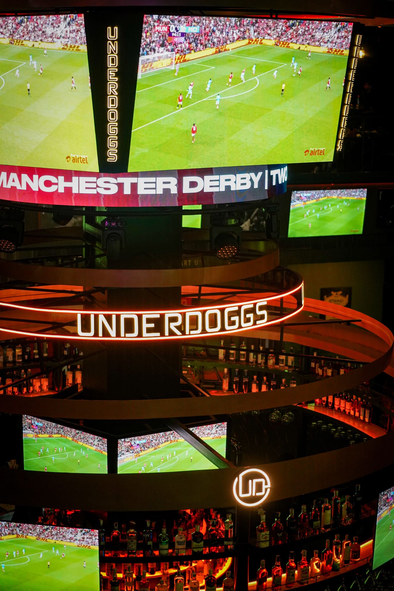 Screens showing a Manchester derby football match above a ring-shaped UNDERDOGGS sign and a lit bar.