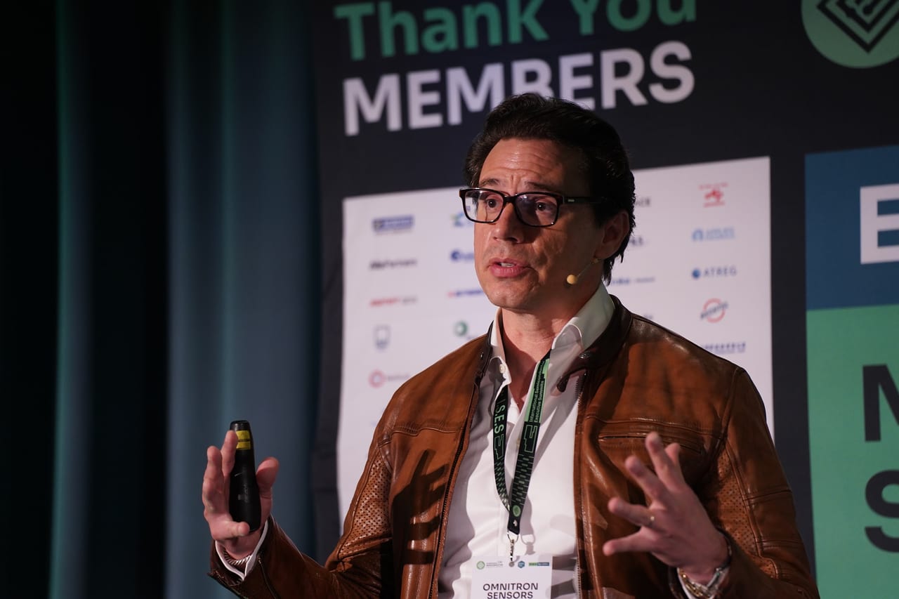 A speaker mid-gesture in front of the members wall at the MEMS World Summit, Porto