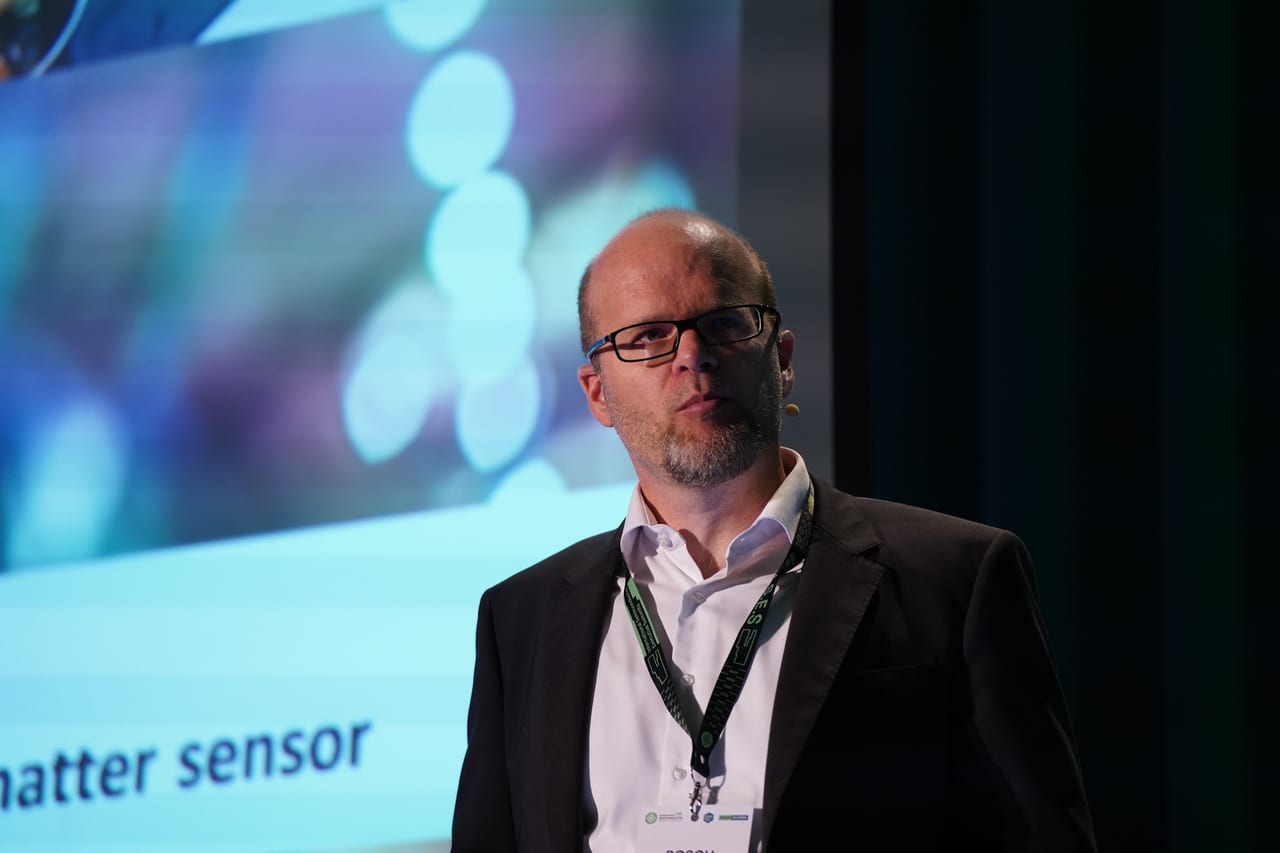 A speaker in profile against a blurred projection at the MEMS World Summit, Porto