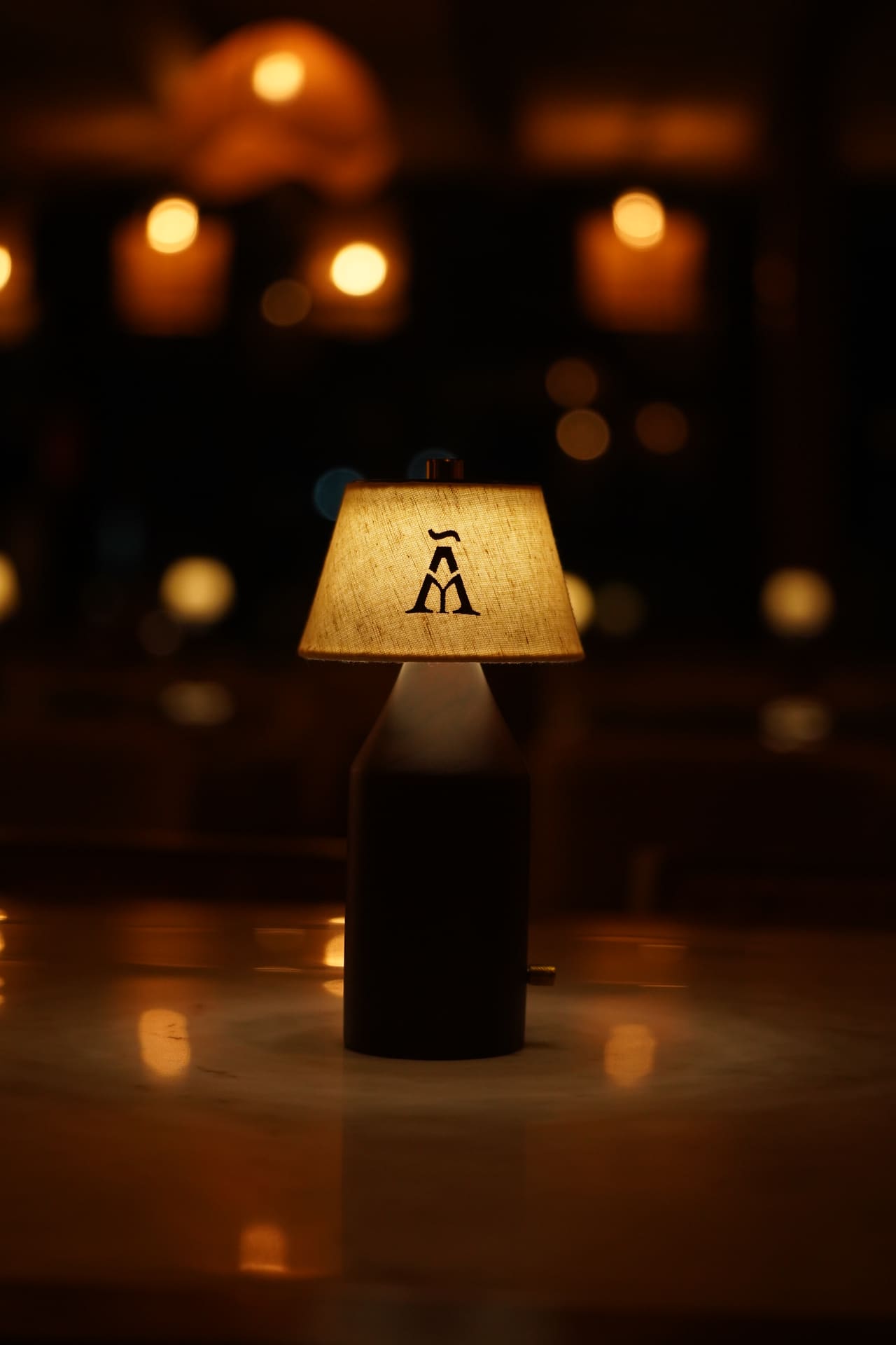 A table lamp with the Ālaya monogram on its shade, glowing on a dark table with warm lights behind.