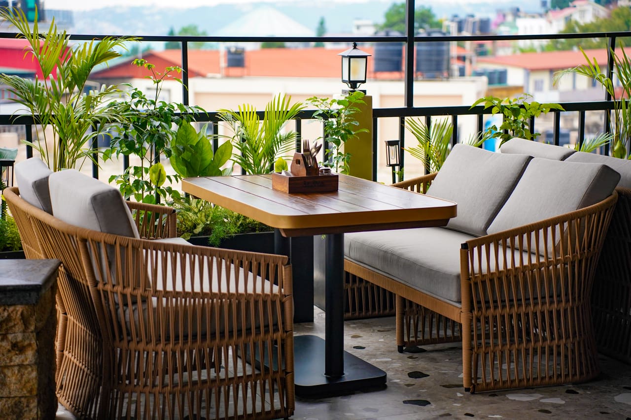 A terrace corner at Ālaya by day: rattan sofas around a timber table, planters and a view over hillside rooftops.