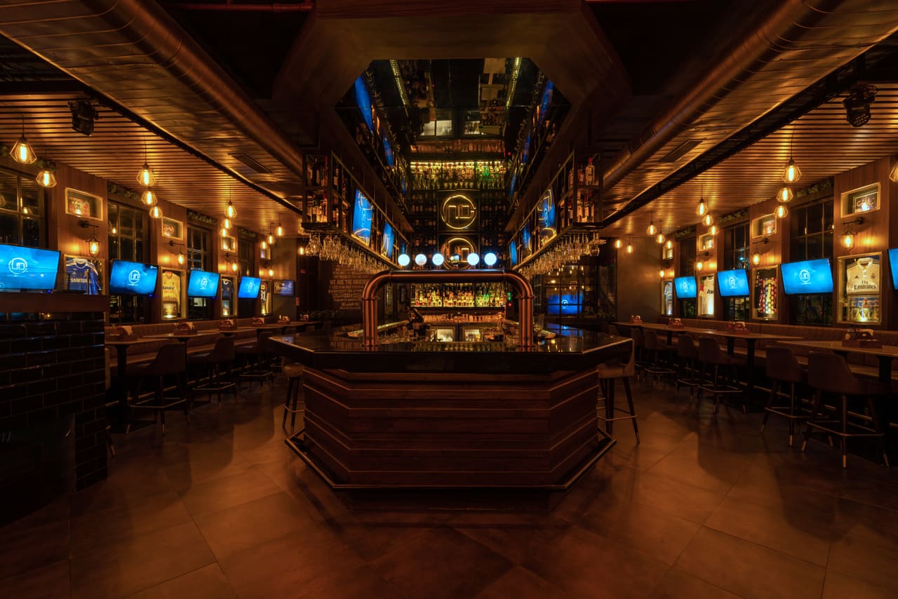 The central island bar at Underdoggs Aerocity under a slatted timber ceiling, with screens along both walls.