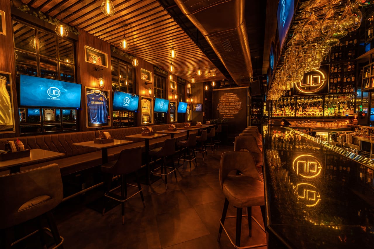 The long bar at Underdoggs Aerocity, its backlit shelves and the UD logo reflected in the polished bar top.